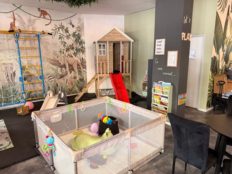 Indoor play area with a large playpen, wooden playhouse structure, red slide, and climbing ladder against nature-themed wall murals