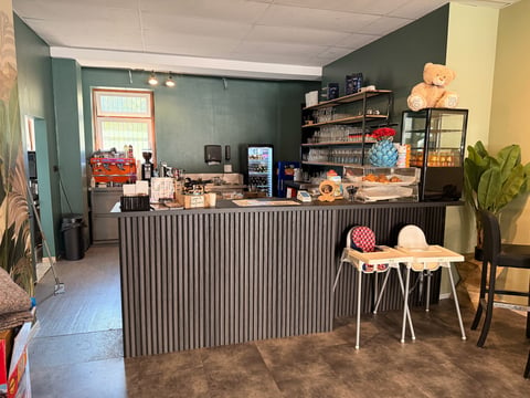 Modern retail or cafe counter with teal walls, corrugated metal desk, shelving with products and toys, small seating area with chairs
