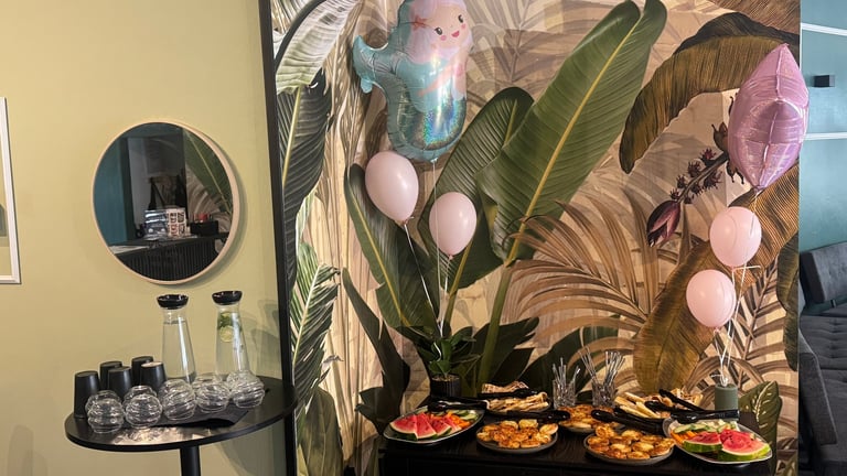 Party setup with tropical themed backdrop, pink balloons, appetizer trays, ring light, and bottles on a black table