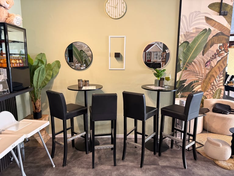 Modern lounge area with high-top black tables, bar stools, potted plants, wall mirrors, and botanical wall art on cream walls