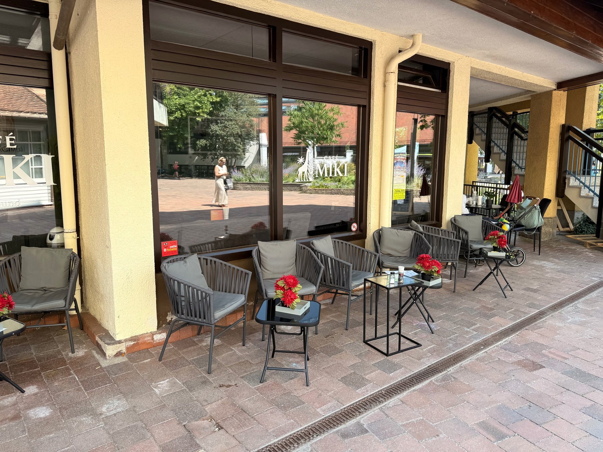 Outdoor covered patio seating area with modern chairs, small tables, and red flowers under a cream and wood awning