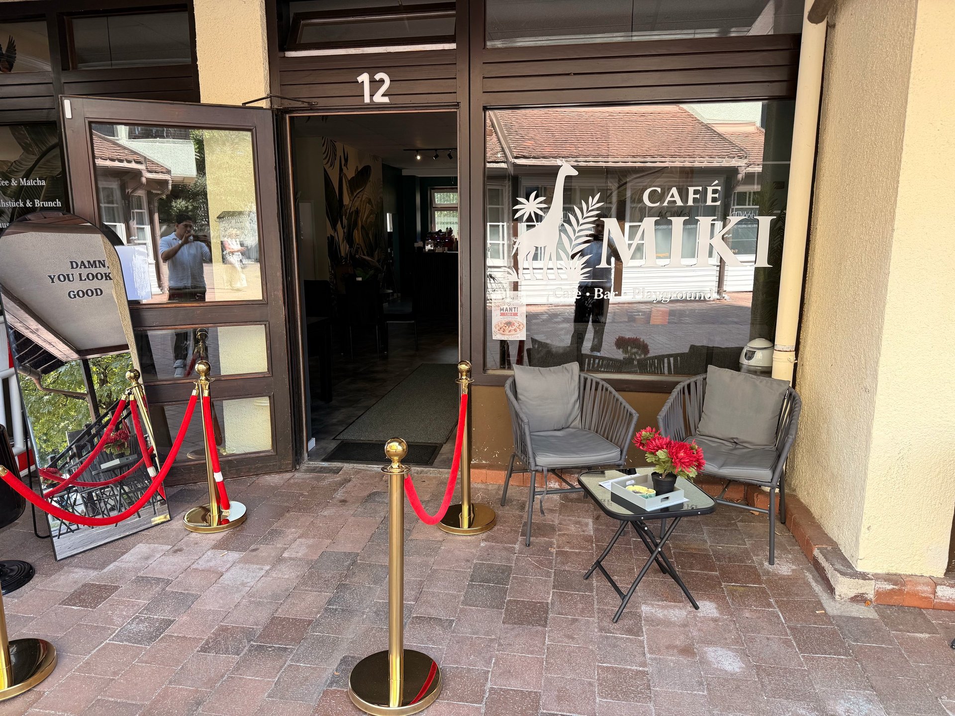 Storefront entrance with red and brass stanchions, white café signage in window, and small black table with flower on terracotta tile floor